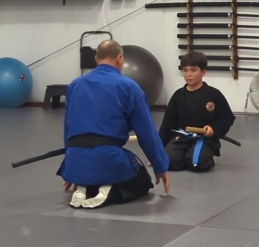 Two martial artists kneeling on a mat, facing each other in a dojo; one in a blue gi, the other in a black gi with a blue belt, holding a wooden sword.