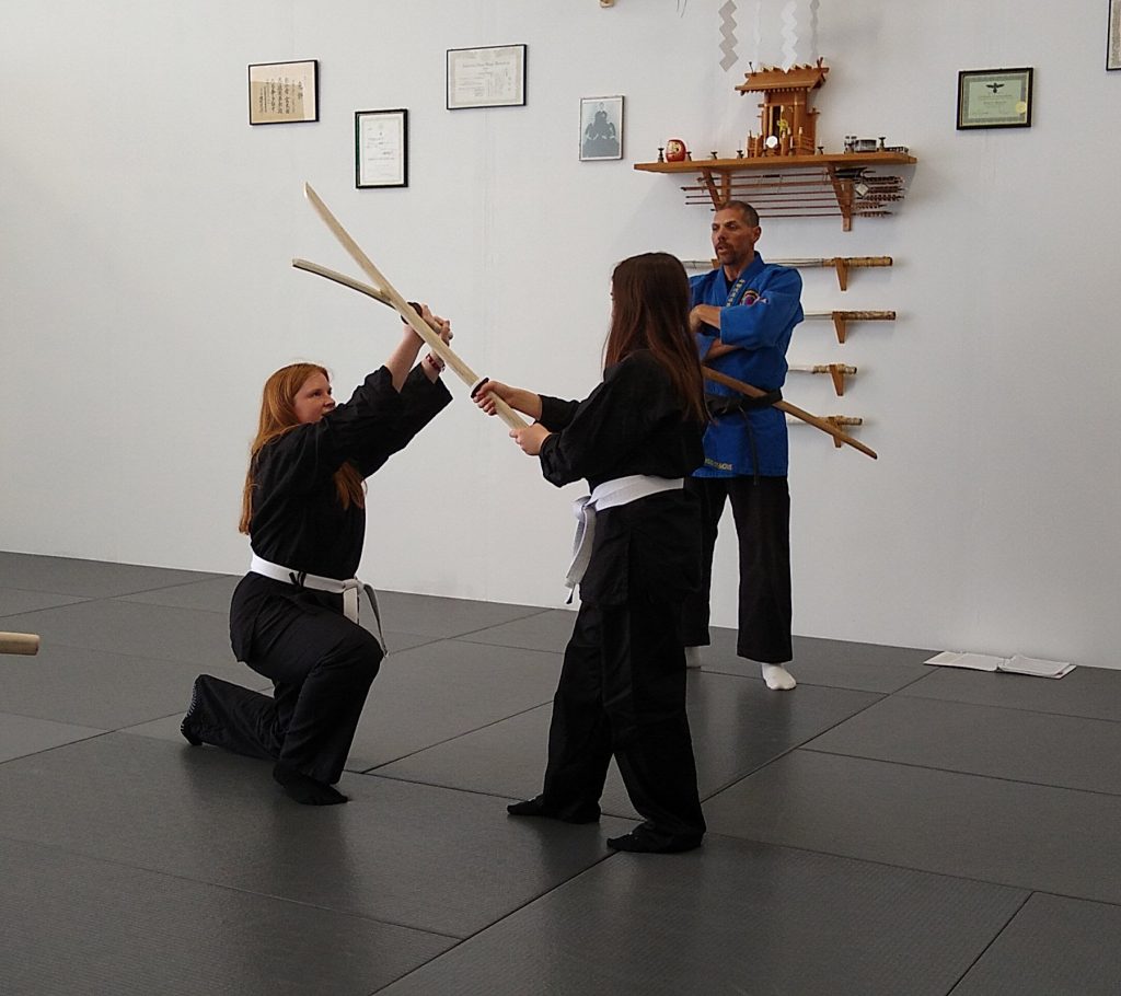 Two martial arts students spar with wooden swords as a blue-gi instructor watches in a dojo with sword rack and certificates on the wall, mats on the floor.