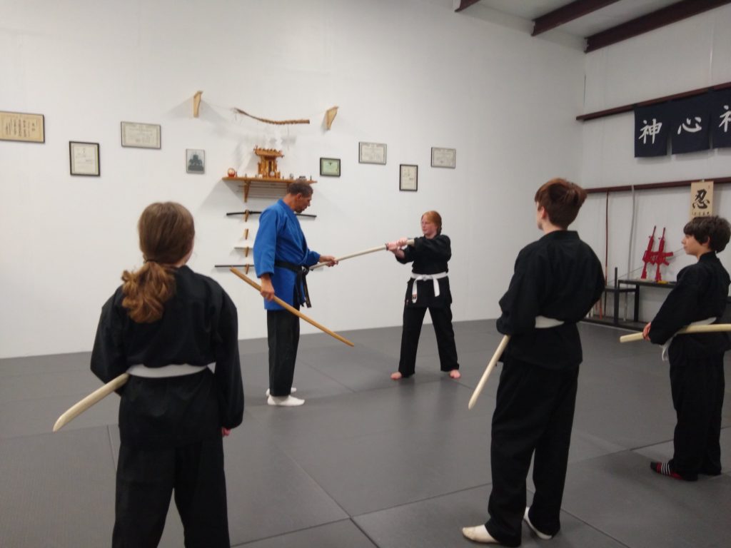 Martial arts class practicing with wooden swords in a dojo; instructor in a blue gi demonstrates to students in black uniforms.