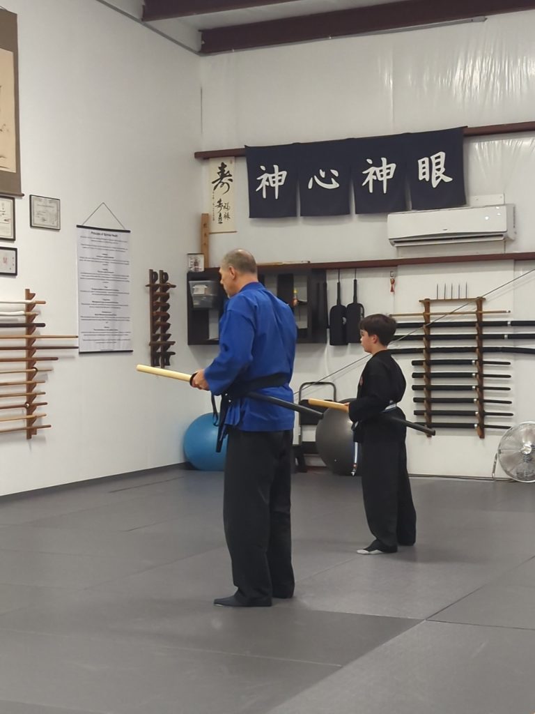 Instructor in a blue gi and a student in a black gi practice with wooden swords in a dojo, kanji banners on the wall above them