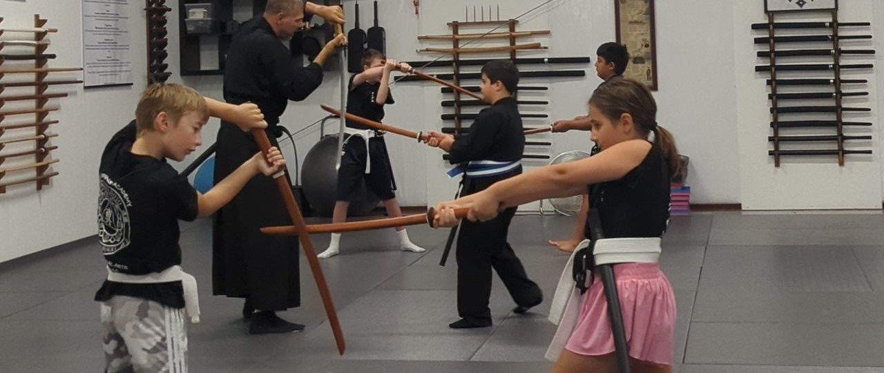 Group of children and instructor practicing swordforms in a martial arts dojo with wooden training swords on padded mats.