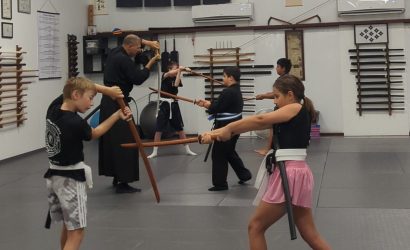 Group of children and instructor practicing swordforms in a martial arts dojo with wooden training swords on padded mats.